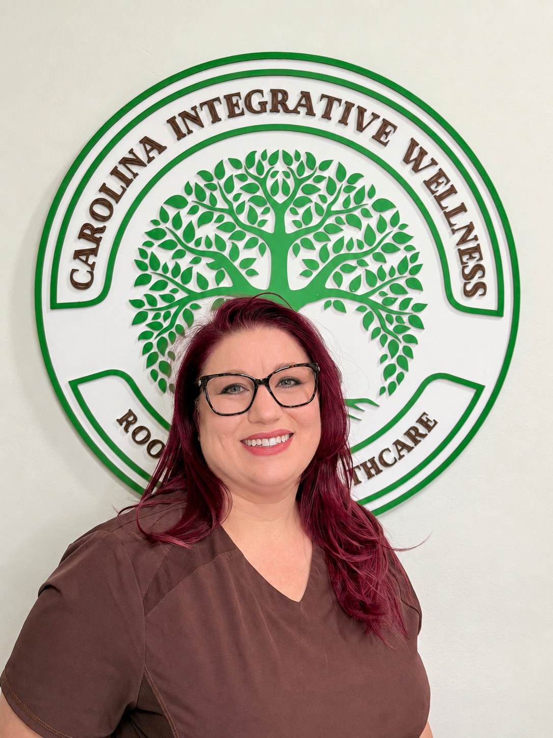 Smiling woman with dark red hair and glasses standing in front of a circular green-and-white sign featuring a tree logo and the text Carolina Integrative Wellness.
